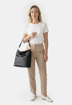 Naine valges t-särgis ja beežides pükstes, hoides käes musta nahast käekotti kuldsete detailidega, valged loafersid jalas, seisab ühevärvilise tausta ees.