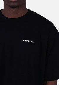 Černé bavlněné tričko s krátkými rukávy a kulatým výstřihem. Slovo "CRYSTAL" je vytištěno bílou barvou na levé straně hrudi.