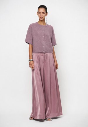 Vrouw die een mauve gekleurde kortemouwige gebuttonede trui en wijde satijnen mauve broek draagt, staand tegen een effen witte achtergrond.