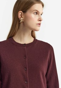 Bordeauxfärgad cardigan med rund halsringning, knäppning och strukturerad stickning. Kombination med örhängen i guldton med textur. Slät tygfinish.