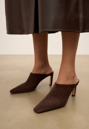 Vrouw draagt donkerbruine, puntige hoge hak mules met een midi-lange bruine leren rok, staand op een gladde beige vloer.