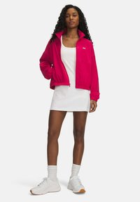 Chaqueta de cremallera fucsia con cuello alto y puños elásticos, llevada sobre un vestido blanco. Modelo de pie, mostrando zapatillas deportivas blancas y calcetines.