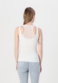 Top a canotta bianca a coste con un design aderente, caratterizzato da pannelli posteriori trasparenti e spalline larghe. Abbinato a jeans azzurri chiari.