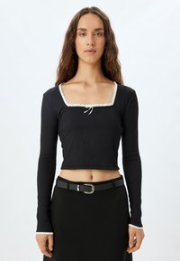 Zwart langemouwen crop top met een vierkante halslijn, voorzien van een witte ruffelrand en een strikdetail aan de voorkant. Gevoelige stof.