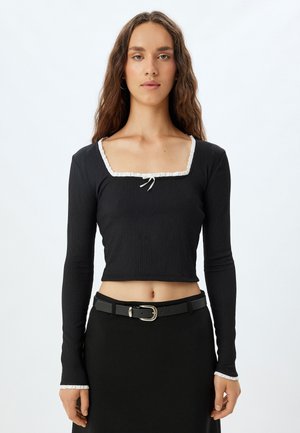 Schwarzes Langarm-Crop-Top mit quadratischem Ausschnitt, verziert mit einer weißen Rüscheneinfassung und einem Frontknotendetail. Texturiertes Material.