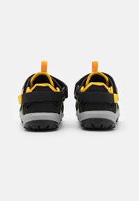 Mustat ja keltaiset lasten kengät, joissa on hengittävä verkkomateriaali, kumipohja, säädettävä remmi ja vetotappi, sekä kestävä rakenne.