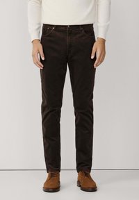 Hombre que lleva pantalones de pana marrón oscuro, camiseta blanca de manga larga y zapatos de ante beige con cordones, de pie contra un fondo liso.
