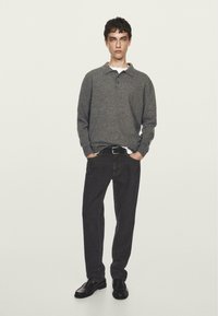 Maglione a polo grigio, jeans scuri e scarpe nere. Il modello sta in piedi con le mani in tasca, indossando una T-shirt bianca sotto.