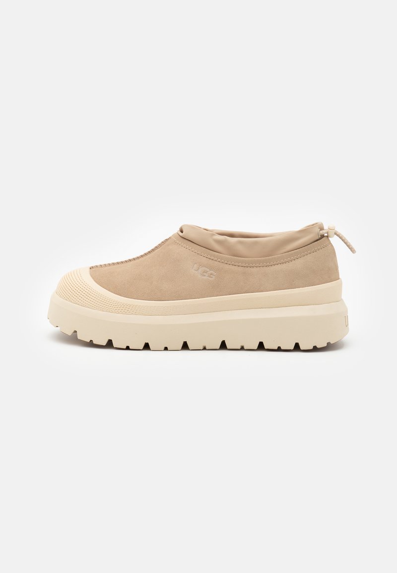 Beige Wildleder Slipper mit gepolstertem Kragen, dicker cremefarbener Gummisohle und dezentem Logodetail an der Seite. Strukturierte Zehenpartie und Trittmuster.
