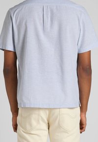 Camicia azzurra a maniche corte con una piega sul retro, realizzata in un tessuto testurizzato. Abbinata a pantaloni beige chiaro con tasche cucite.