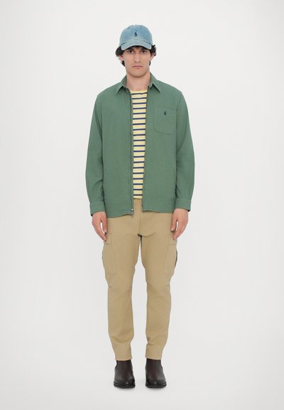 Camisa verde con cremallera, camiseta a rayas amarilla y azul marino, pantalones cargo beige y una gorra de mezclilla clara, complementados por botas negras.