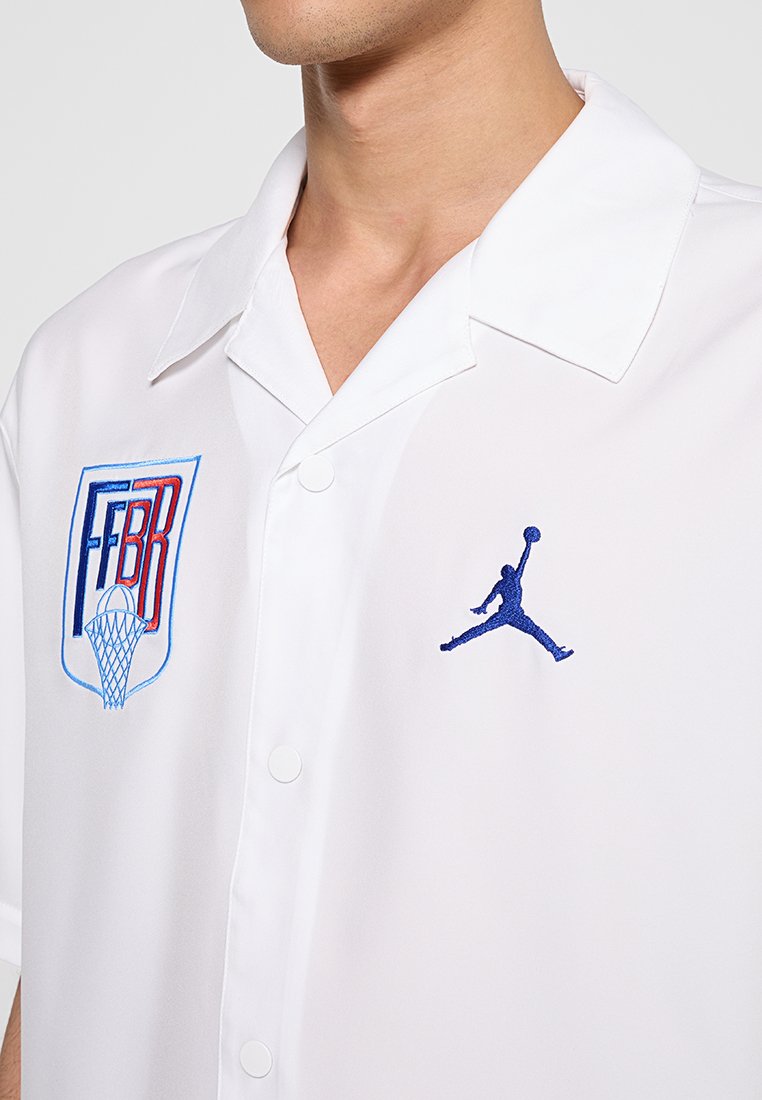Camisa corta de manga blanca con logotipos de baloncesto bordados en azul y rojo en el lado izquierdo del pecho y un logotipo de Jumpman en azul en el lado derecho.