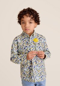 Lange mouwen knoopshirt van lichte stof, met een meerkleurig bladpatroon. Een kind houdt een gele bloem in de hand en is gekleed in lichtblauwe jeans.