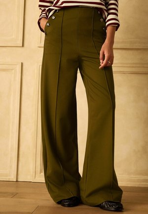 Olivgrüne, weit geschnittene Hose mit hoher Taille, vertikalen Nähten und metallischen Knöpfen, getragen mit schwarzen Schuhen und einem gestreiften Langarmoberteil.