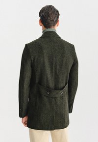Ciemnozielony wełniany płaszcz o teksturowanym wykończeniu, z podwójnym zapięciem na guziki z tyłu i dopasowanym krojem, widocznymi liniami szwów oraz kołnierzem z wycięciami.