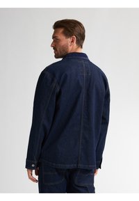 Mørk denimjakke med krave og lange ærmer, synlig syning og knapdetaljer. Stoffet har et struktureret, robust udseende.