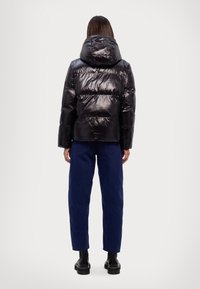 Veste matelassée noire brillante avec capuche, dotée de sections matelassées et d'une coupe décontractée, portée avec un pantalon bleu et des bottes noires.