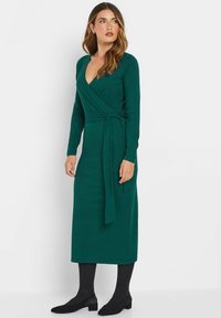 Langärmliges Wickelkleid in Dunkelgrün mit V-Ausschnitt, Bindegürtel und glatter Textur, kombiniert mit schwarzen Strumpfhosen und flachen Schuhen mit niedrigen Absätzen.