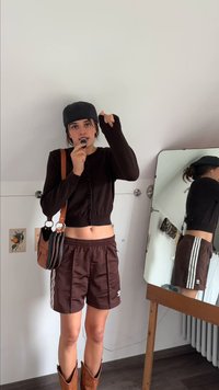 Brązowy, krótki kardigan, brązowe spodenki sportowe z białymi paskami, brązowe skórzane botki, szara czapka i brązowa torebka na ramię. Lustro w tle.