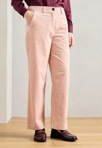 Osoba stoji obučena u svijetlosmeđe kord pantalone, bordo majicu dugih rukava i sjajne bordo mokasine na drvenom podu.