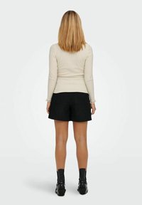 Kobieta z blond włosami do ramion, ubrana w kremowy top z długim rękawem, czarne teksturowane szorty i czarne botki za kostkę, stojąca tyłem.