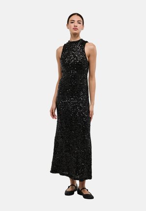 Robe de soirée noire à sequins avec un col montant et un design sans manches, s'étirant jusqu'au sol. Assortie à des chaussures plates noires scintillantes.