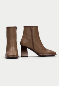 Botas de piel marrón hasta el tobillo con puntera puntiaguda, cierre lateral con cremallera y tacón cuadrado apilado. Textura suave con un diseño minimalista.
