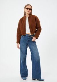 Giacca bomber in pile marrone con chiusura a zip, top bianco corto e jeans blu a gamba larga. Gli accessori includono occhiali da sole scuri e orecchini.
