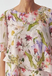Blouse à motif floral en tissu transparent. Présente des fleurs de différentes couleurs et des feuilles vertes sur un fond rose clair. Encolure ronde, coupe ample.