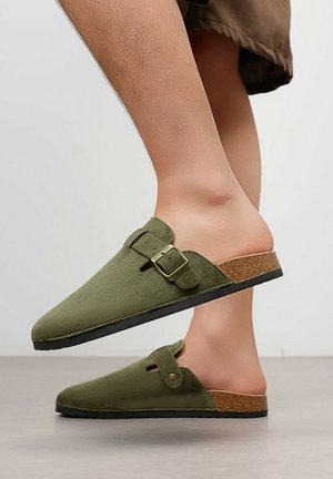 Personne portant des sabots en daim vert olive à enfiler avec des brides à boucle ajustable et des semelles en liège, debout sur un sol lisse.