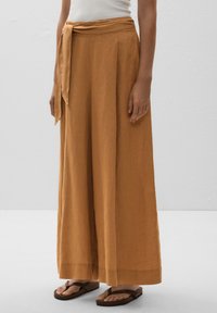 Wide-leg trousers in a burnt orange linen fabric, featuring a tied waist and a relaxed silhouette. Paired with brown flip-flops.