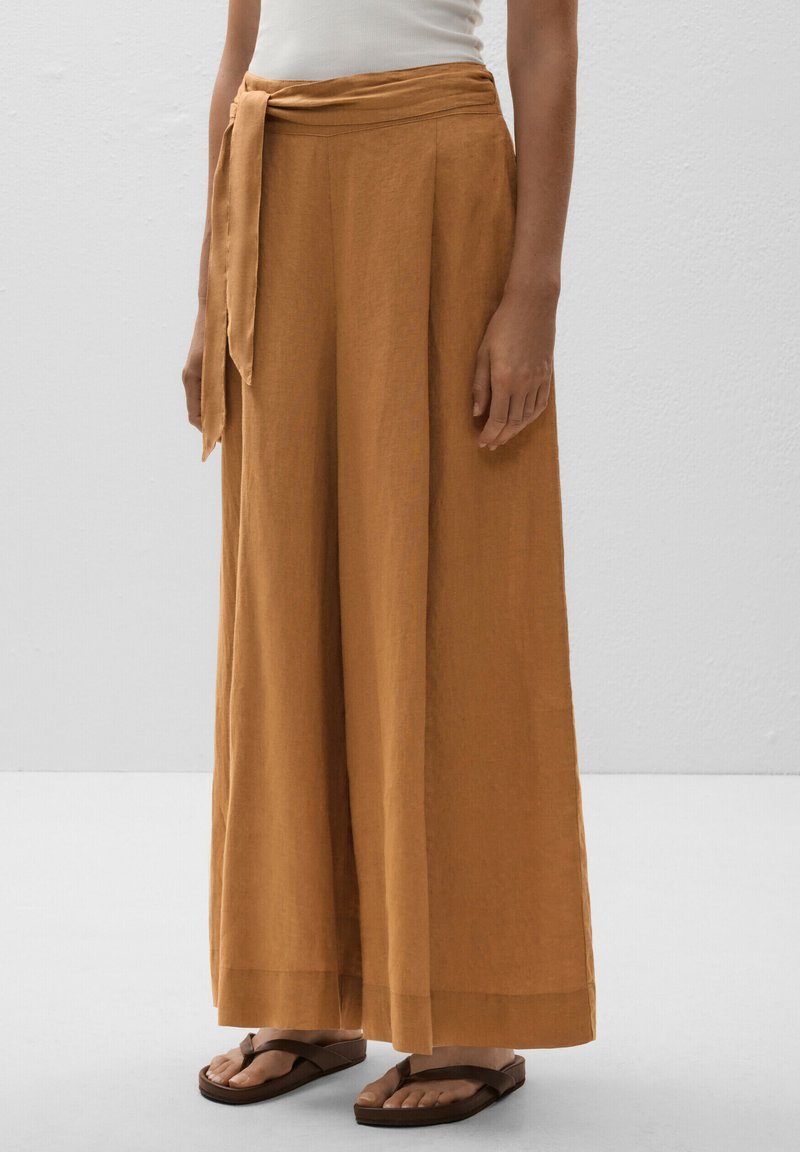 Wide-leg trousers in a burnt orange linen fabric, featuring a tied waist and a relaxed silhouette. Paired with brown flip-flops.
