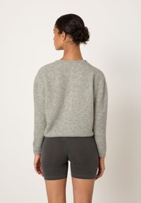 Žena s tmavými vlasy svázanými do drdolu, která má zlaté kruhové náušnice, šedý chlupatý svetr a tmavě šedé přiléhavé šortky, stojící před jednobarevným pozadím.