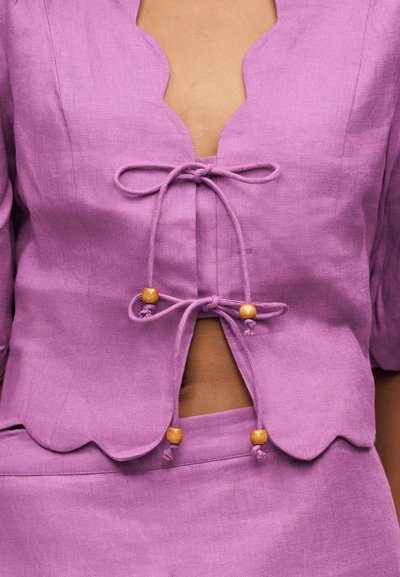 Haut en lin violet avec bords festonnés et liens décoratifs ornés de perles en bois, partiellement ouvert à l'avant, porté avec une jupe ou un pantalon assorti.