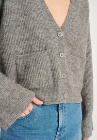 Grau gerippter Cardigan mit tiefem V-Ausschnitt, der vier Knöpfe und zwei Fronttaschen hat. Aus weichem Material gefertigt, kombiniert mit blauen Jeans.