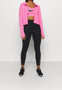 Roze sportieve zip-up hoodie, cropped, met lange mouwen boven een bijpassende roze sportbeha. Zwarte high-waisted leggings met zij-uitsnijdingen. Zwarte sneakers.