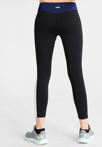 Ben som bär svarta leggings med vita sidosträck och grå sportskor med mintgröna sulor, står på en ljus bakgrund.