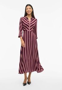 Langes, knopfbares Kleid in burgunderroten und hellrosa Streifen. Hat einen Kragen und Dreiviertelärmel, mit einem fließenden Rock und kombiniert mit schwarzen High Heels.