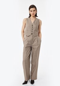 Brauner Leinenweste mit fünf schwarzen Knöpfen, kombiniert mit hochgeschnittenen, weiten Hosen im gleichen Stoff. Stilettos runden den Look ab.