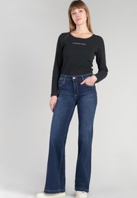 Jeans de tiro medio en azul oscuro con campana, fabricados en denim. El modelo lleva un top negro de manga larga con textura ribeteada y detalles en texto.