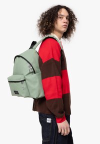 Mochila verde con detalles negros, que presenta un bolsillo frontal con cremallera y correas ajustables, llevada por una persona con cabello rizado y un suéter a rayas.