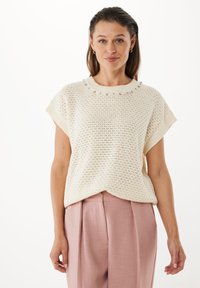Hellescremefarbener gestrickter Pullover mit kurzen Ärmeln, der ein strukturiertes Muster und dekorative Perlen am Rundhalsausschnitt aufweist. Kombiniert mit rosa Hosen.
