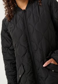 Černá prošívaná bunda s předním zipem, žebrovanými manžetami a kapsou se zapínáním na patentek. Materiál má hladkou, jemnou texturu.