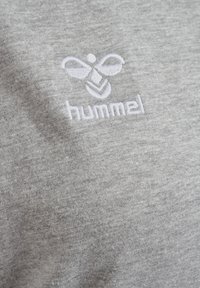 Hvit brodert hummel-logo med stilisert bie og tekst på lys grått stoff, sannsynligvis et nærbilde av et klesplagg.