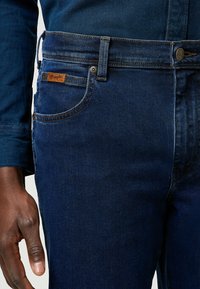 Dark blue denim jeans, featuring a classic cut, a front pocket, and a leather patch on the waistband with brand logo. Smooth texture.