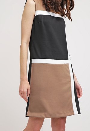 Femme portant une robe sans manches bicolore noire et marron avec une bordure blanche, debout devant un fond neutre uni.