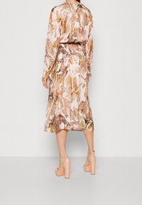 Abito a maniche lunghe con un motivo marmorizzato rosa, marrone e oro. La vita è cintrata e l'abito è abbinato a scarpe con tacco nude.