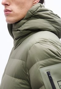 Κλείσιμο εικόνας ενός ατόμου που φοράει πράσινο μπουφάν με επένδυση, ψηλό γιακά και φερμουάρ σε τσέπη στο μανίκι.