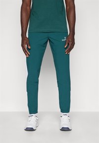 Teal joggerbukser med hvide sidelommer, elastiske manchetter og et logo. Kombineret med en mørkegrøn T-shirt og hvide sportsko.