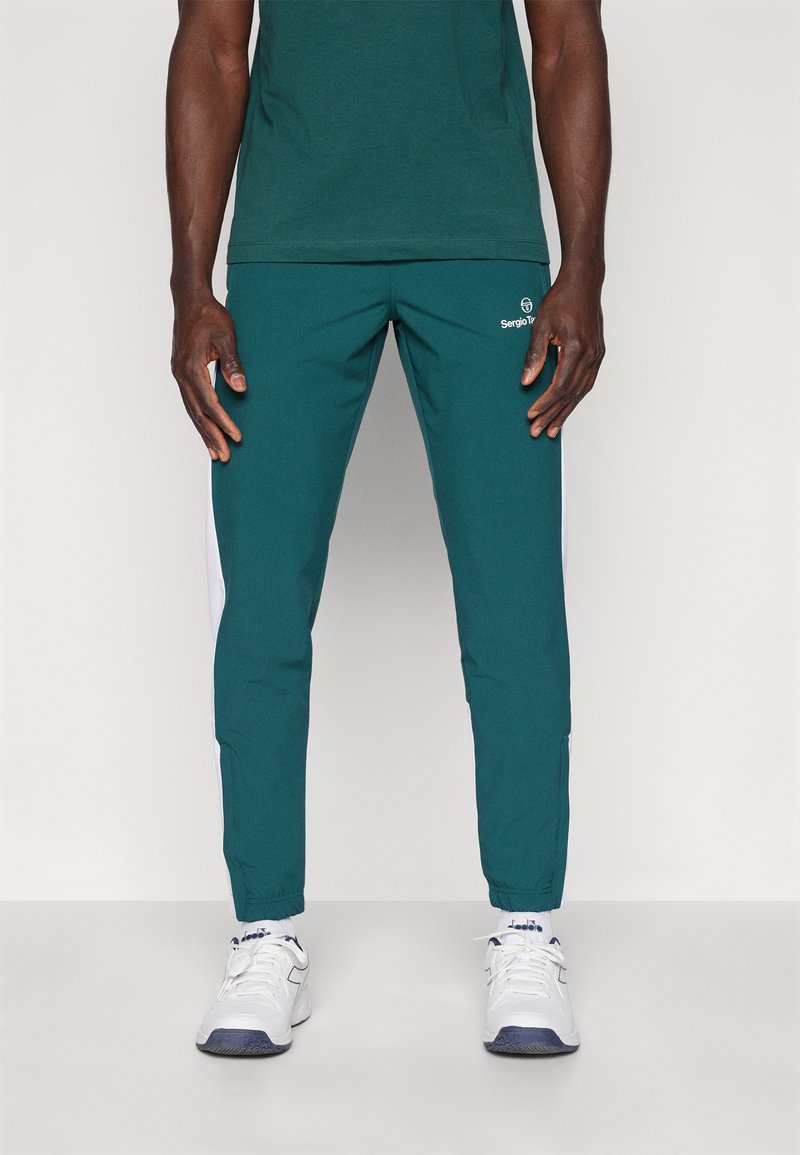 Teal joggerbukser med hvide sidelommer, elastiske manchetter og et logo. Kombineret med en mørkegrøn T-shirt og hvide sportsko.